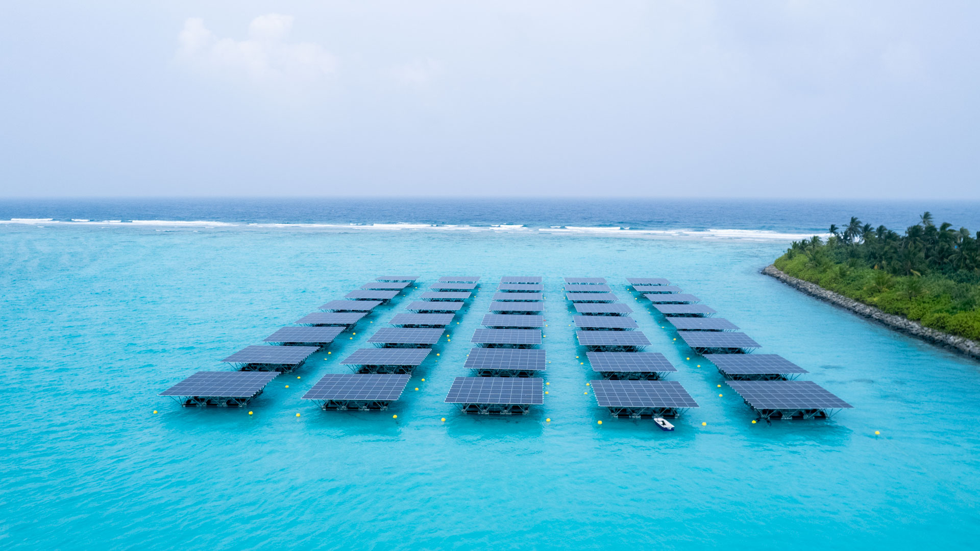 Aerial view of a floating solar PV array in a bright turquoise Maldivian lagoon, with five parallel rows of blue solar panels on elevated aluminum platforms, yellow mooring buoys around the perimeter, a small service boat alongside, and a palm-covered island shoreline to the right with a reef break and open ocean in the distance.